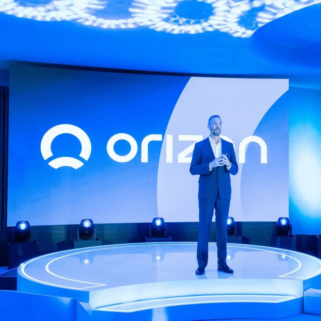 Παρουσίαση της Orizon σε εταιρική εκδήλωση με ομιλητή στη σκηνή και φωτισμένο λογότυπο στο φόντο.